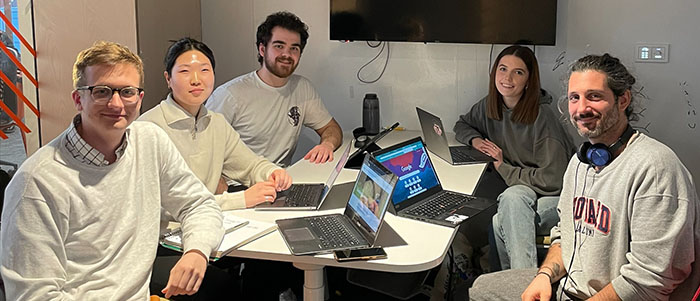 Five students sitting around a desk using laptops