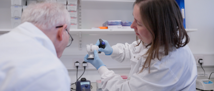 Female wearing lab coat and blue gloves, holding genomics lab equipment and showing a male. 