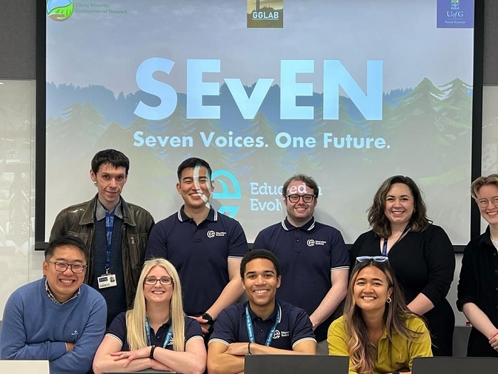 10 team members smiling for the camera wiht a screen behind them displaying 'SEvEN. Seven Voices. One Future.'