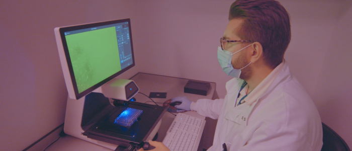 Male person wearing facemask and lab coat sitting in front of a screen in a room lit by red lights.