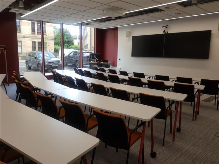 Flat floored teaching room with rows of tables and chairs, two large screens, videoconferencing camera, PC, and lectern.