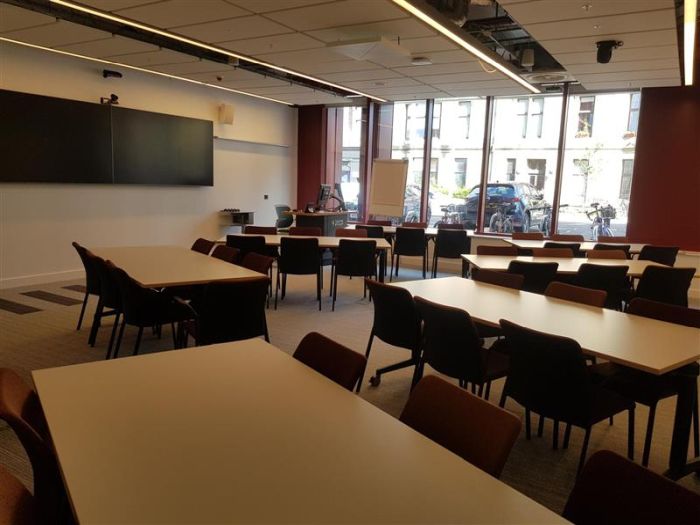 Flat floored teaching room with tables and chairs in groups, two large video monitors, videoconferencing camera, PC, and lectern.