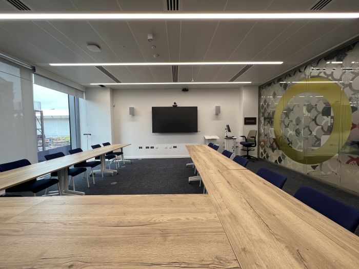 Flat-floored meeting room with tables and chairs in a horseshoe layout, wall-mounted video monitor, videoconferencing camera, speakers and microphone, and lectern.