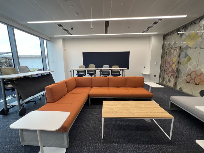 Flat-floored meeting room with high tables and chairs and sofa arranged in parallel L-shape layout, coffee table, side table, and low padded bench.