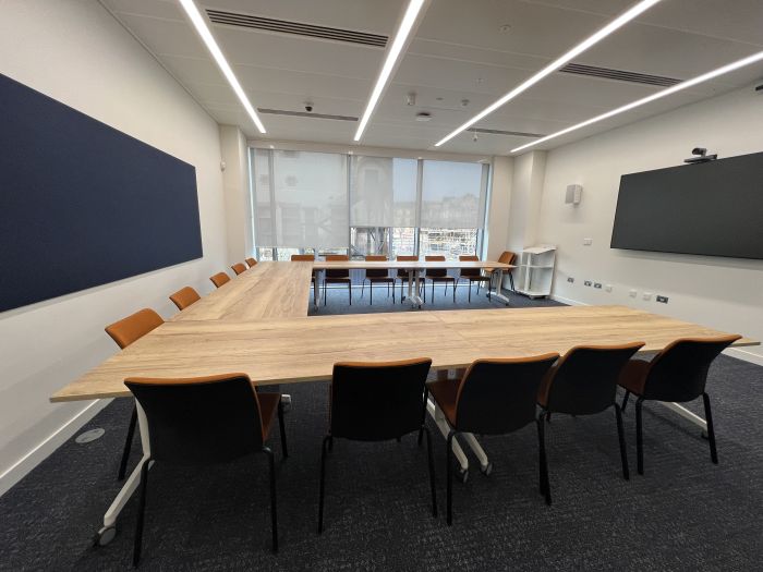 Flat-floored meeting room with tables and chairs in a horseshoe layout, wall mounted video monitor, videoconferencing camera and speakers, and lectern.