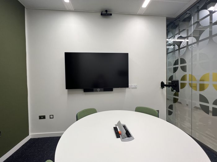 Flat-floored meeting room with round table and chairs, and wall mounted video monitor.