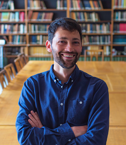 Image of Daniel Streicker in a blue shirt sitting on a desk with his arms folded and smiling