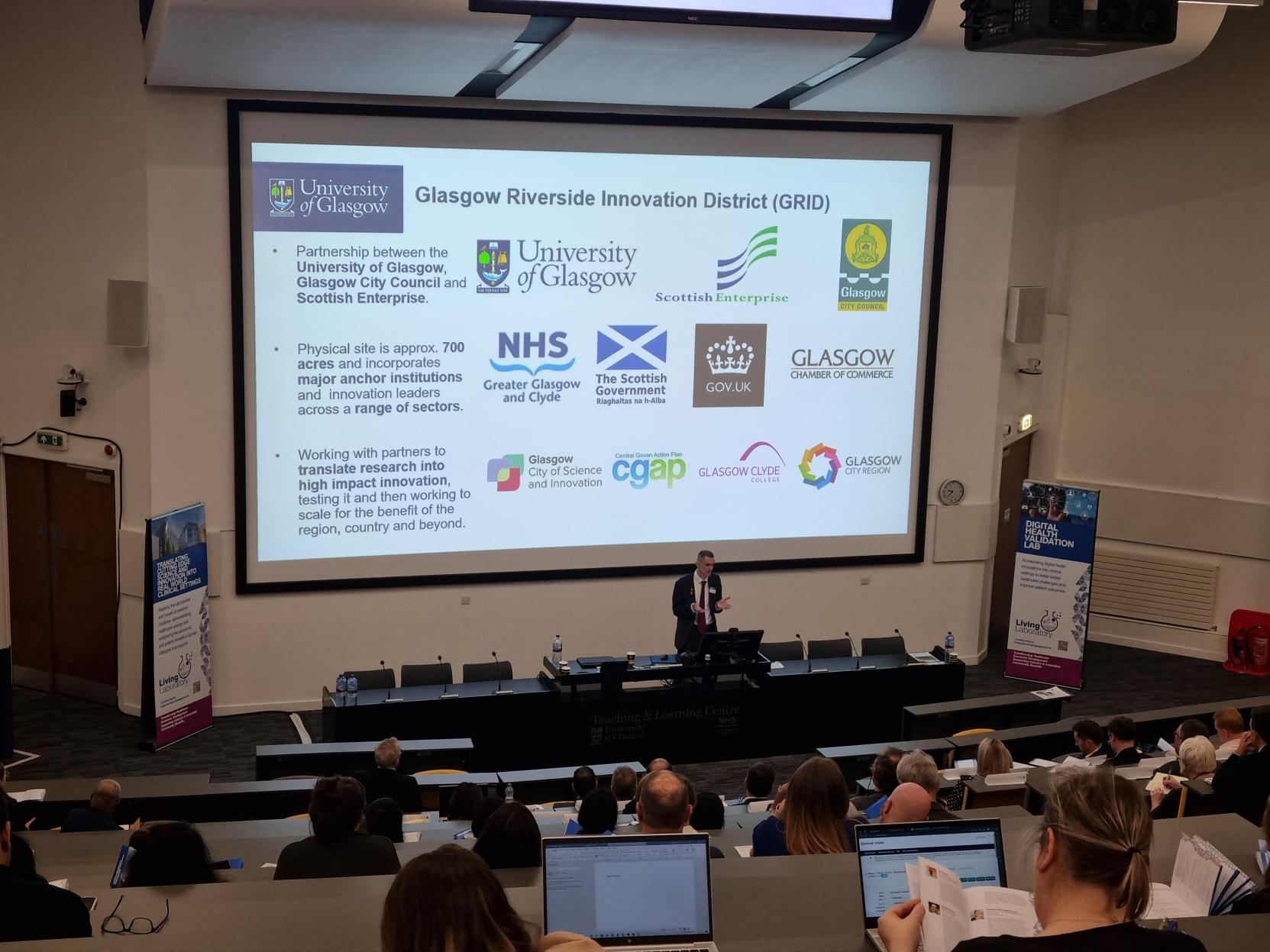 Mike King delivers talk on GRID in front of screen showcasing the partnership between UofG, Scottish Enterprise, Glasgow City Council, NHS GG&C, The Scottish Government, GOV.UK, Glasgow City of Commerce, Glasgow City of Science and Innovation, Central Govan Action Plan, Glasgow Clyde College and Glasgow City Region