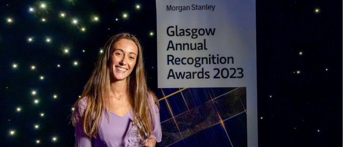 A woman stands holding a trophy before a sign advertising the Glasgow Annual Recognition awards