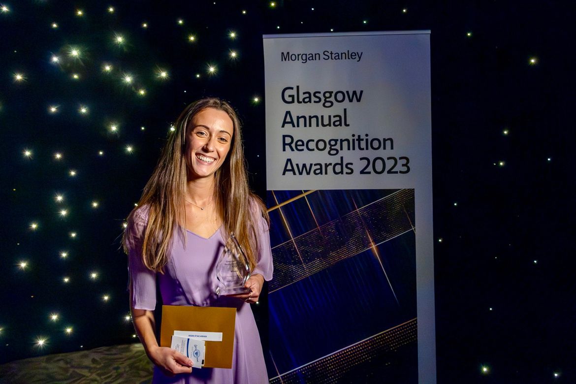A woman stands holding a trophy before a sign advertising the Glasgow Annual Recognition awards