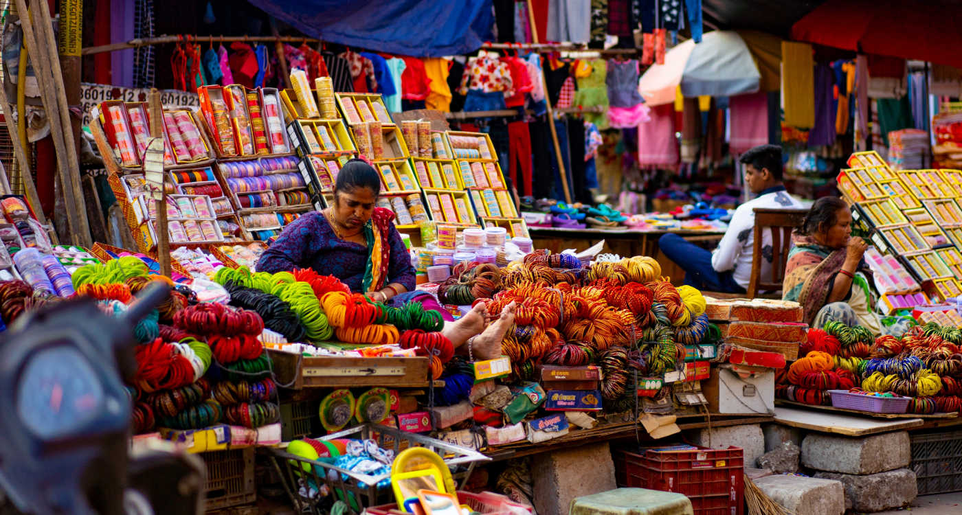 Woman sitting at a marketplace stall selling a very colourful selection of goods such as wool and clothing [Photo: Alin Andersen, Unsplash]
