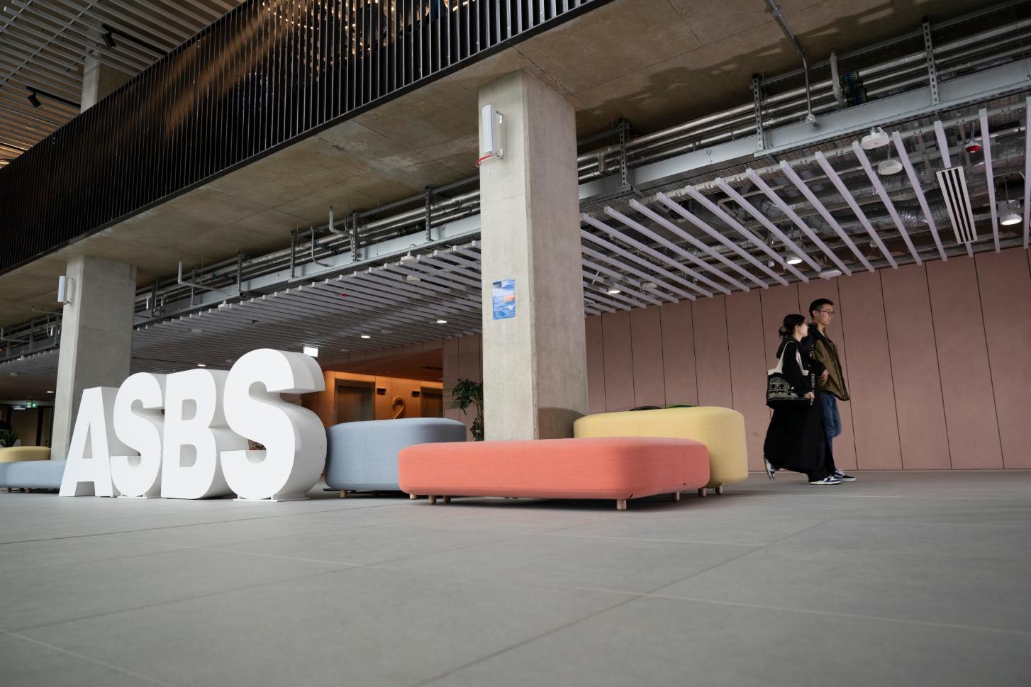 Two people walk through the lobby of the ASBS PG hub. Large white letters spell out ASBS. 