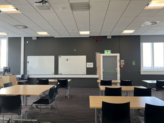 Flat floored teaching room with tables and chairs in rows and groups, whiteboards, lectern, projector, lecturer's chair, and PC.