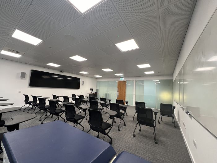 Flat floored teaching room with tablet chairs, clinical skills bed, whiteboards, video monitors, lectern, and PC.