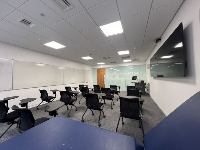 Flat floored teaching room with tablet chairs, clinical skills bed, whiteboards, video monitors, lectern, and PC.