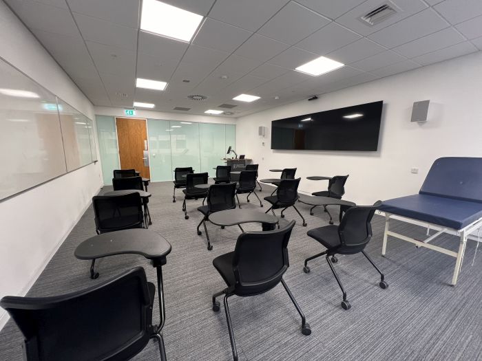 Flat floored teaching room with tablet chairs, clinical skills bed, whiteboards, video monitors, lectern, and PC.