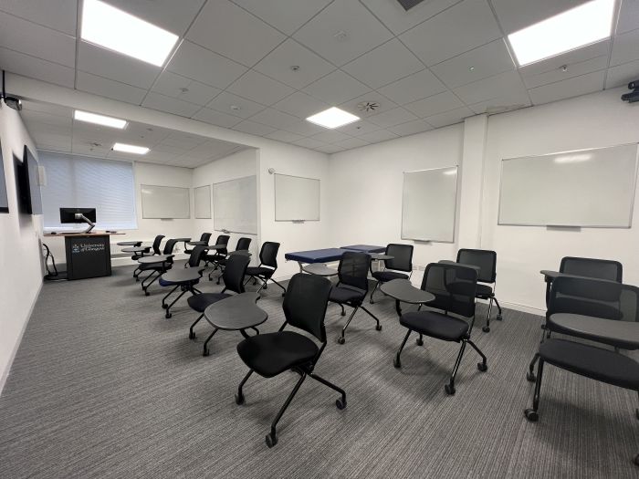Flat floored teaching room with rows of tablet chairs, clinical skills bed, whiteboards, video monitors, lectern, and PC.