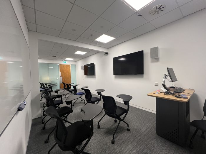 Flat floored teaching room with tablet chairs, whiteboards, video monitors, lectern, and PC.