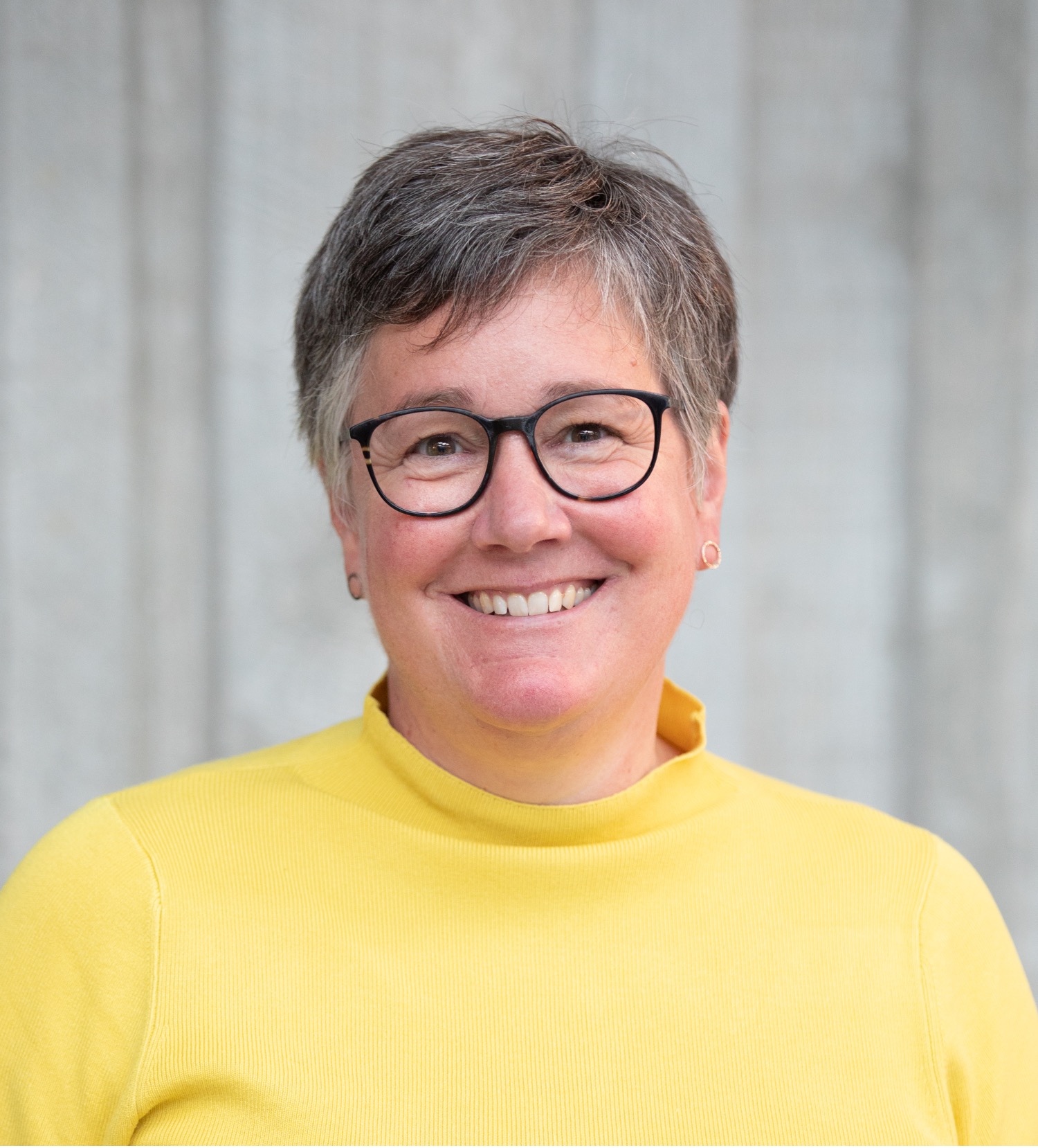 Christina Naula - This photograph shows a headshot of Christina, a pale skinned woman with grey short hair, glasses and a bit smile. She is wearing a yellow jumper