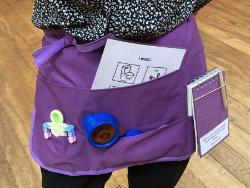 A person wearing a cloth apron with items in pockets: fidget toy; notebook; toy glasses; communication card
