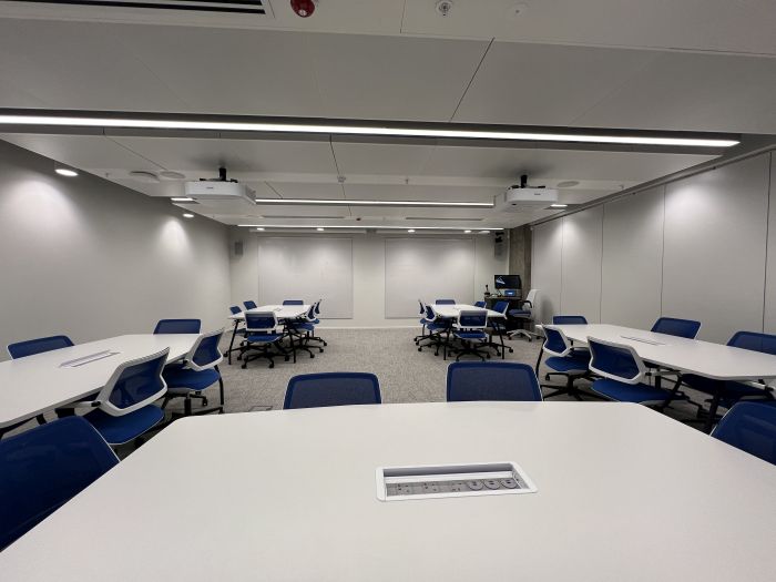 Flat floored teaching room with tables and chairs, projectors, whiteboards, lectern, PC, and lecturer's chair..