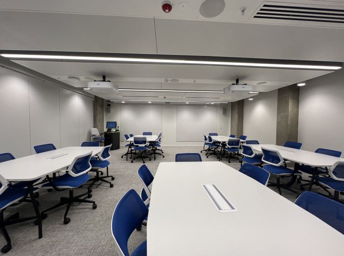 Flat floored teaching room with tables and chairs, projectors, whiteboards, lectern, PC, and lecturer's chair.