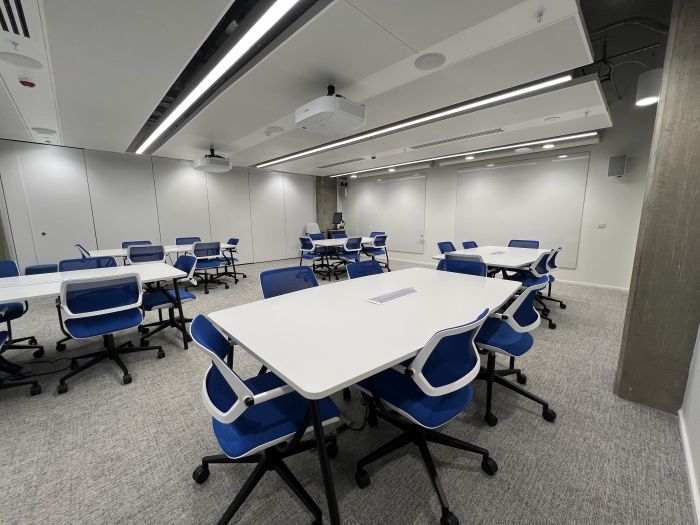 Flat floored teaching room with tables and chairs, projectors, whiteboards, lectern, PC, and lecturer's chair.