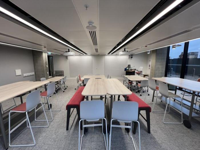Flat floored teaching room with various tables of mixed height, chairs and bench seating, projectors, wall-mounted whiteboards, lectern, PC, and lecturer's chair.