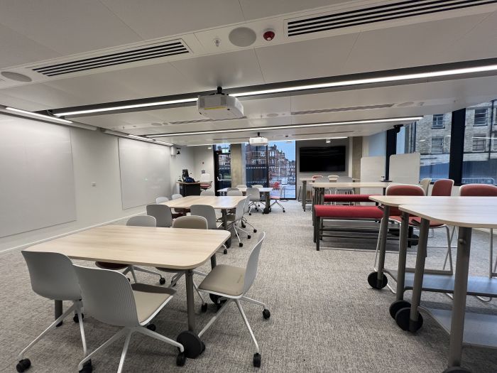 Flat floored teaching room with various tables of mixed height, chairs and bench seating, projectors, wall-mounted whiteboards, moveable whiteboards, large video monitor, lectern, PC, and lecturer's chair.