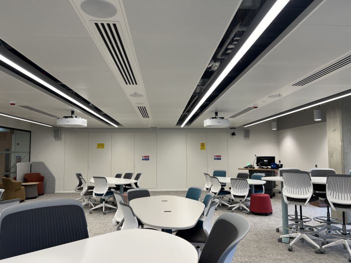 Flat floored teaching room with various tables of mixed height, chairs and stools, projectors, lectern, and PC.