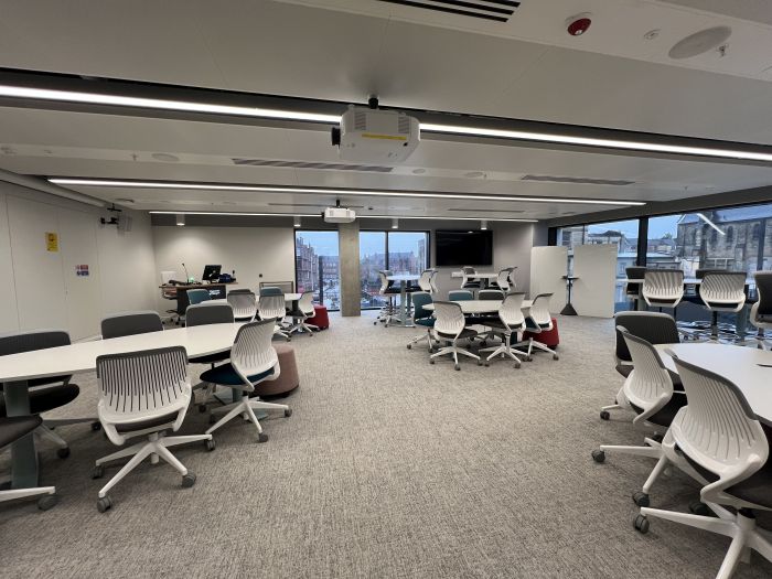 Flat floored teaching room with various tables of mixed height, chairs and stools, projectors, moveable whiteboards, large video monitor, lectern, PC, and lecturer's chair.