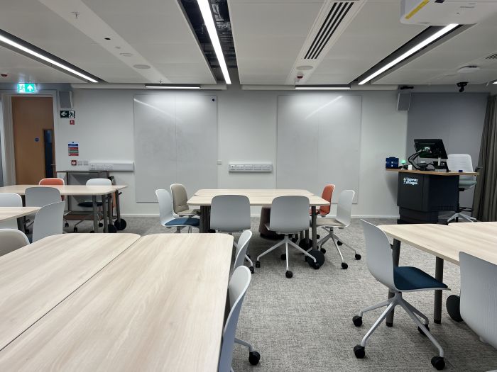 Flat floored teaching room with tables and chairs, projector, whiteboards, lectern, PC, and lecturer's chair.
