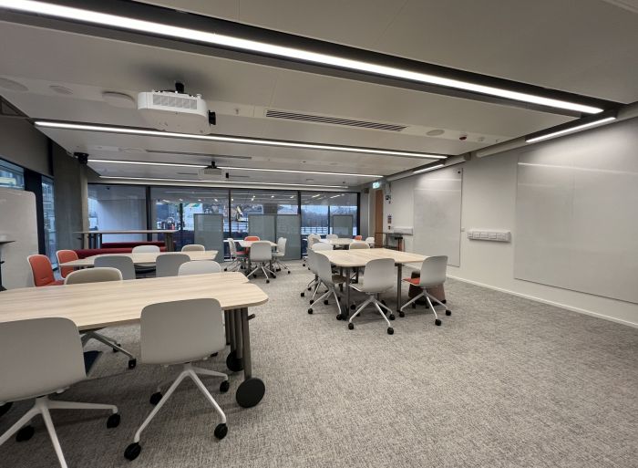 Flat floored teaching room with various tables of mixed height, chairs and bench seating, projectors, wall-mounted whiteboards, and moveable whiteboards.