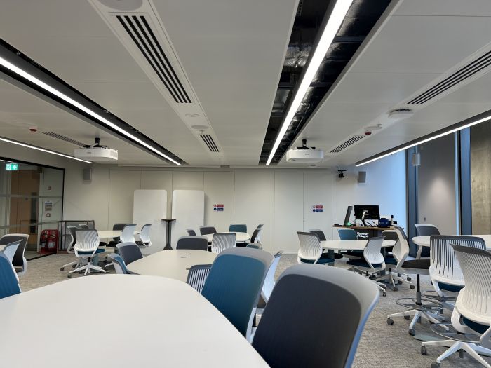 Flat floored teaching room with various tables and chairs of mixed height, projectors, moveable whiteboards, lectern, PC, and lecturer's chair.