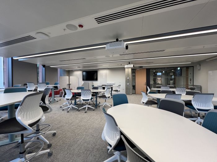 Flat floored teaching room with various tables and chairs of mixed height, projectors, moveable whiteboards, and large video monitor.