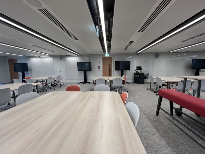 Flat floored teaching room with various tables of mixed height, chairs and bench seating, whiteboards, large video monitors, lectern, PC, and lecturer's chair.