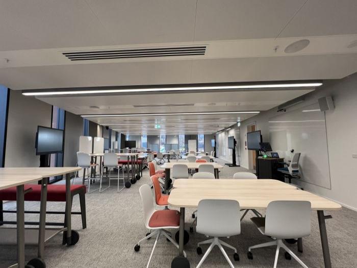 Flat floored teaching room with various tables of mixed height, chairs and bench seating, wall-mounted whiteboards, moveable whiteboards, large video monitors, lectern, PC, and lecturer's chair.