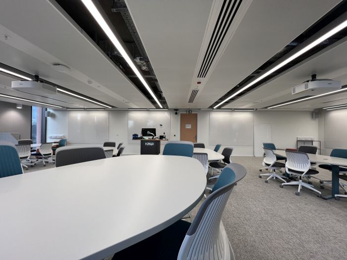 Flat floored teaching room with various tables and chairs of mixed height, projectors, whiteboards, lectern, and PC.