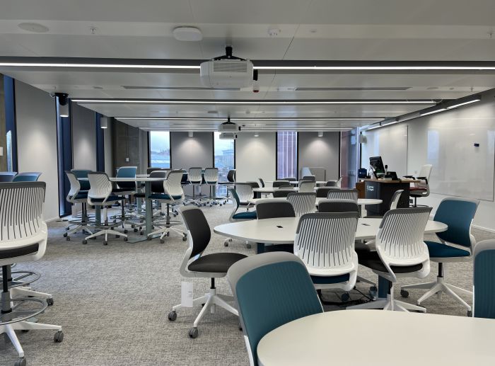 Flat floored teaching room with various tables and chairs of mixed height, projectors, whiteboards, lectern, PC, and lecturer's chair.