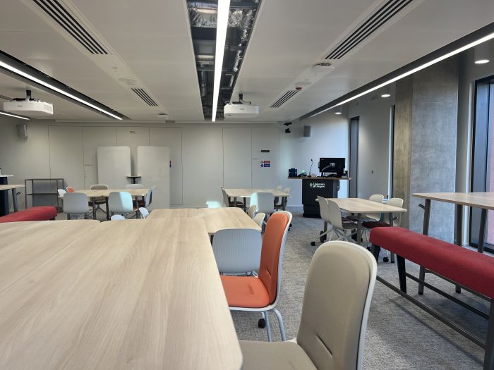 Flat floored teaching room with various tables of mixed height, chairs and bench seating, moveable whiteboards, lectern, and PC.