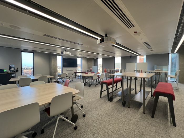 Flat floored teaching room with various tables of mixed height, chairs and bench seating, moveable whiteboards, large video monitor, lectern, and PC.