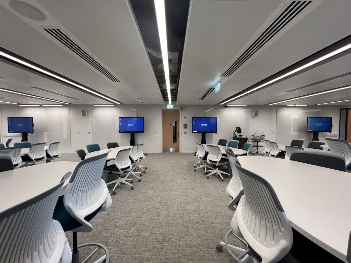 Flat floored teaching room with various tables and chairs of mixed height, whiteboards, large video monitors, lectern, PC, and lecturer's chair.