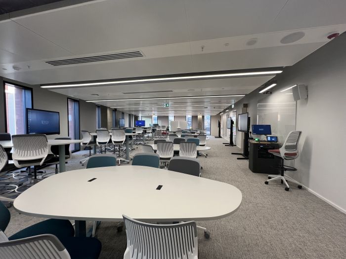 Flat floored teaching room with various tables and chairs of mixed height, whiteboards, large video monitors, lectern, PC, and lecturer's chair.