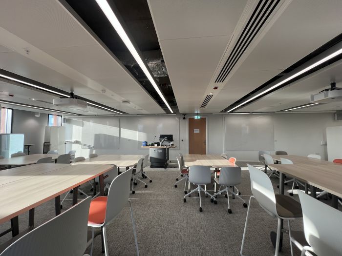 Flat floored teaching room with various tables and chairs of mixed height, projectors, wall-mounted whiteboards, moveable whiteboards,  lectern, and PC.