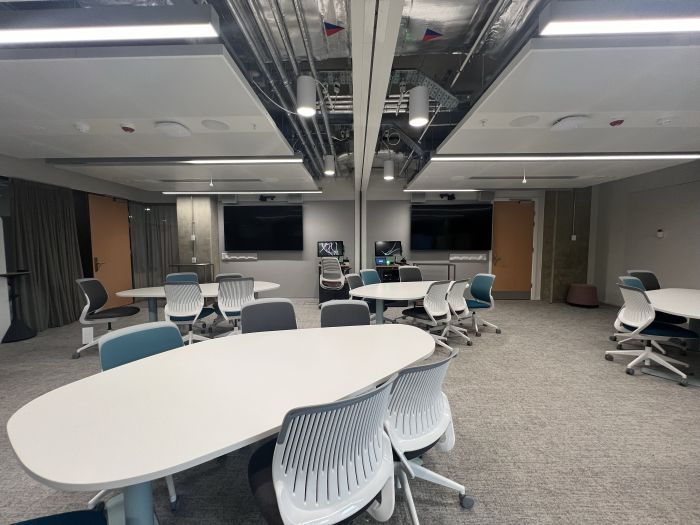 Flat floored teaching room with tables and chairs, moveable whiteboards, large video monitors, lecterns, PCs, and lecturer's chairs.