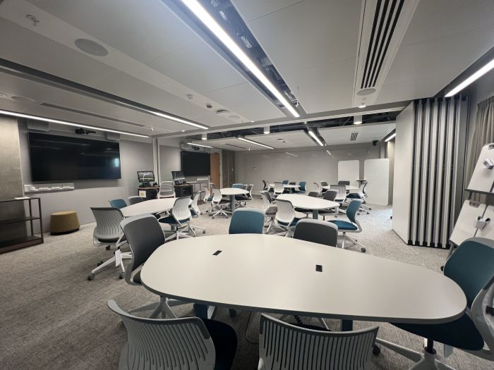 Flat floored teaching room with tables and chairs, moveable whiteboards, large video monitors, lecterns, PCs, and lecturer's chairs.