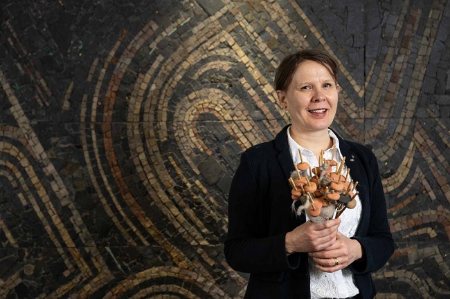  Dr Susanna Harris standing in front of a swirly mosaic holding a bunch of round ceramic weights on sticks. These are replica spindles, tools used to twist fibres into yarn.
