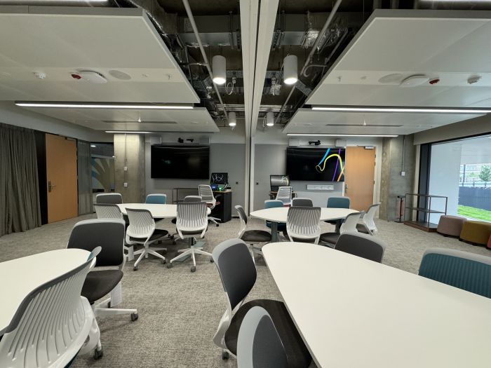 Flat floored teaching room with tables and chairs, large video monitors, lecterns, PCs, and lecturer's chairs.
