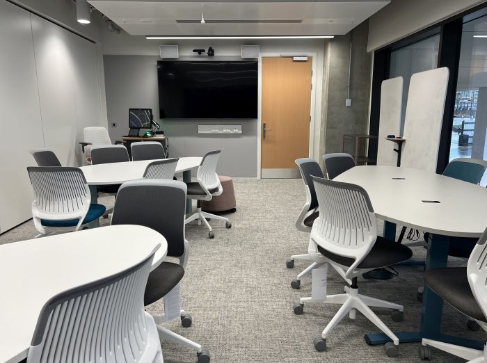 Flat floored teaching room with tables and chairs, moveable whiteboards, large video monitor, lectern, PC, and lecturer's chair. 