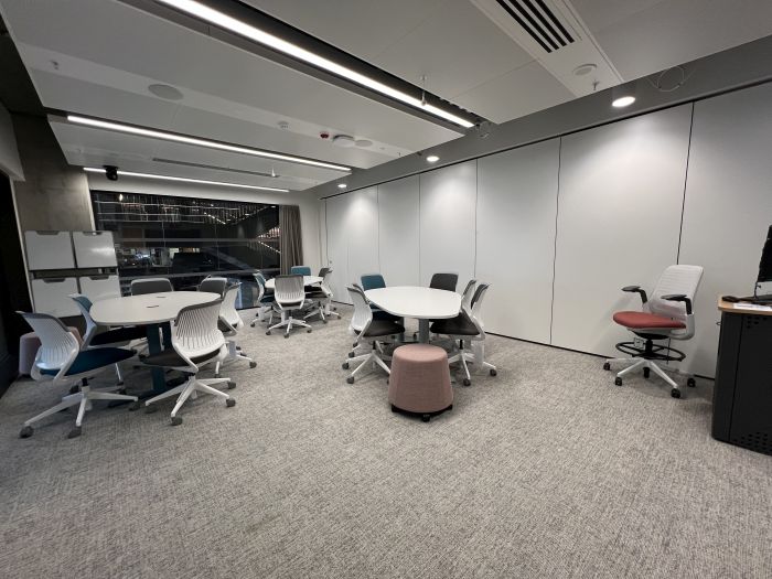 Flat floored teaching room with tables and chairs, stools, moveable whiteboards, lectern, and lecturer's chair. 